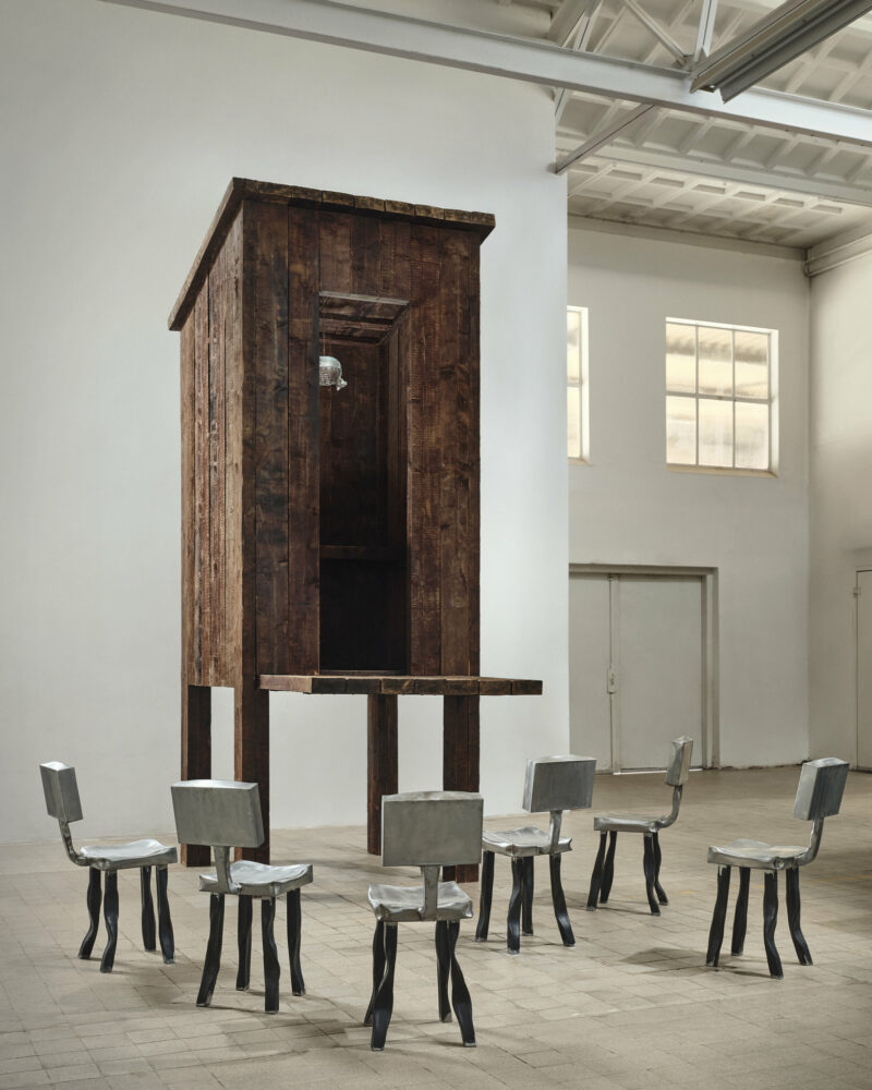 A tall, wooden structure resembling an outdoor privy on stilts, surrounded by six metallic chairs arranged in a circle.