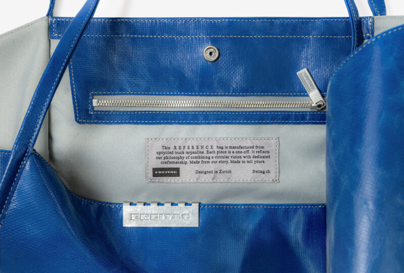Interior of a blue recycled truck tarpaulin Freitag Reference collection tote bag, showing a zippered pocket and a brand information patch.