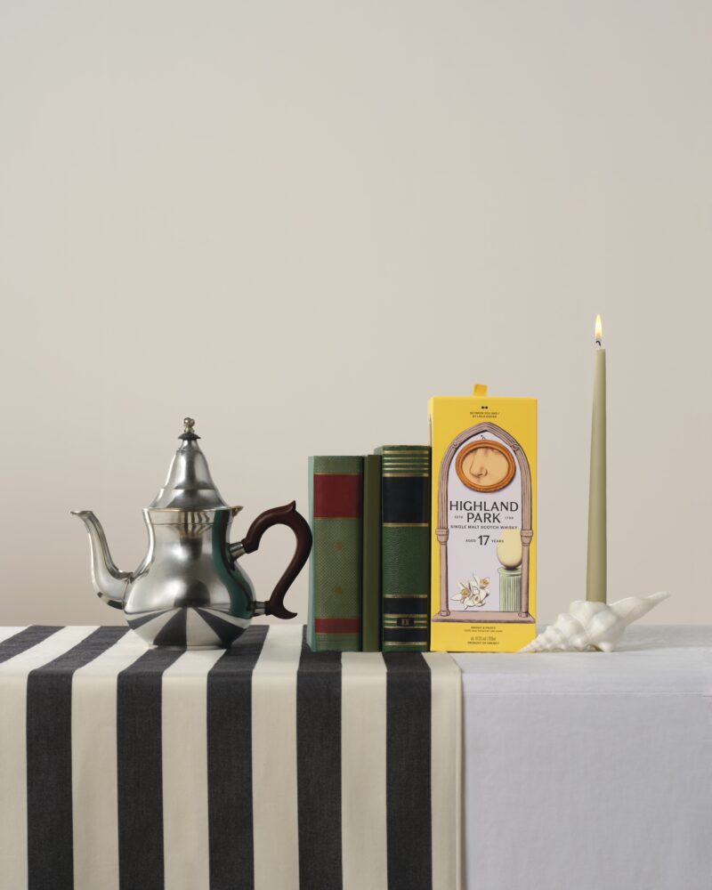 A silver teapot, two closed books, a box labeled “Highland Park 17,” a green candle in a seashell holder, and a striped tablecloth are arranged on a table.