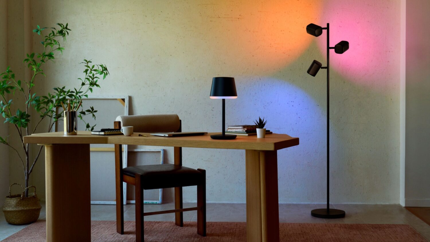 A wooden desk with a lamp and office supplies, next to a Govee floor lamp casting orange, pink, and blue light on the wall.