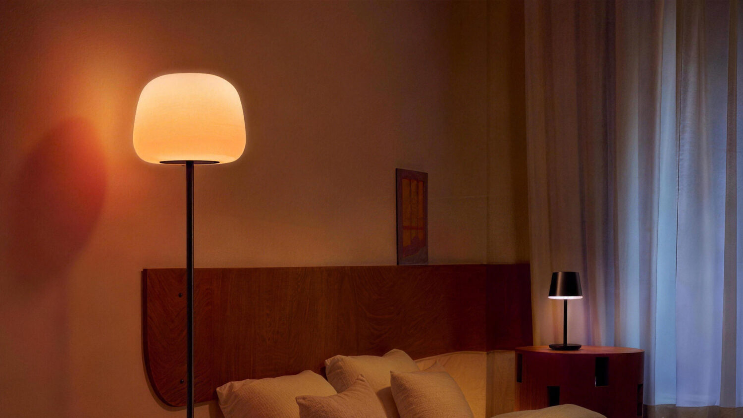 A bedroom interior featuring a tall Govee floor lamp with a glowing, bowl-shaped shade and a small table lamp on a bedside stand.