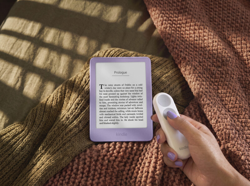 A hand holds a remote next to a purple Kindle redesign e-reader displaying a page of text, resting on folded blankets.