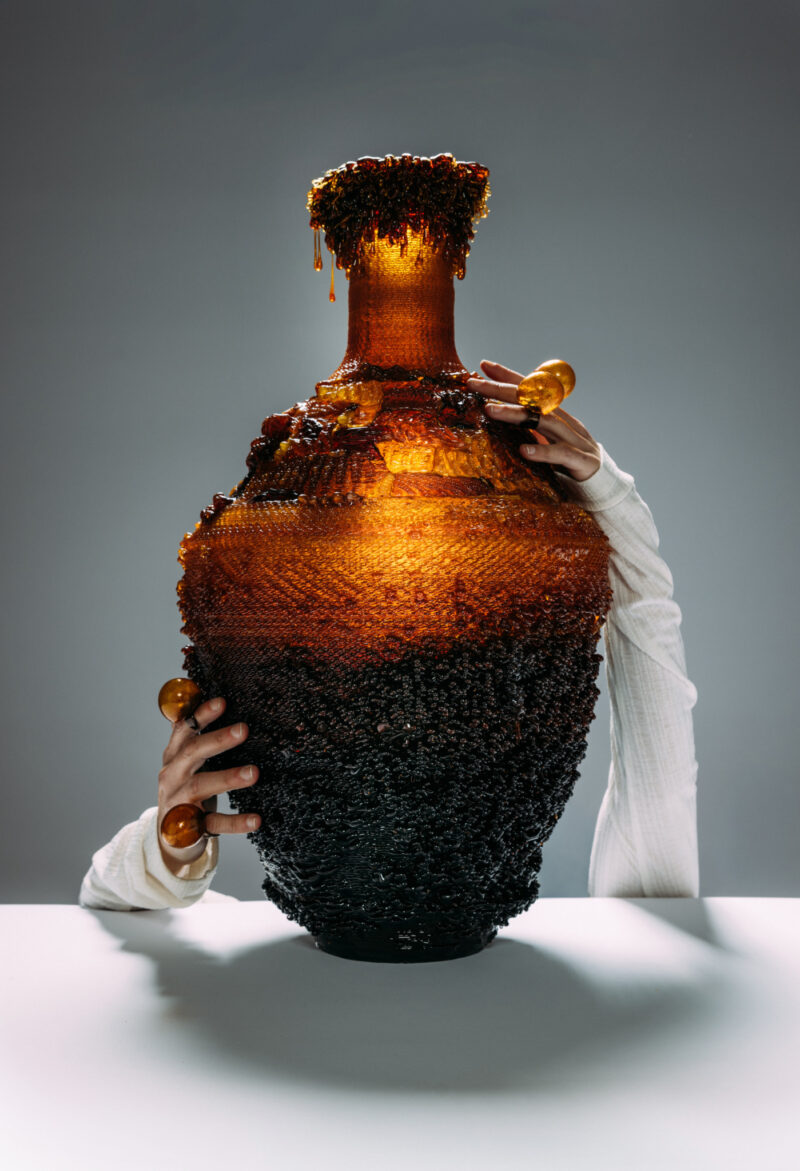A person in a white long-sleeved shirt holds a large, textured vase that transitions from black at the base to amber at the top, where it appears to be dripping.