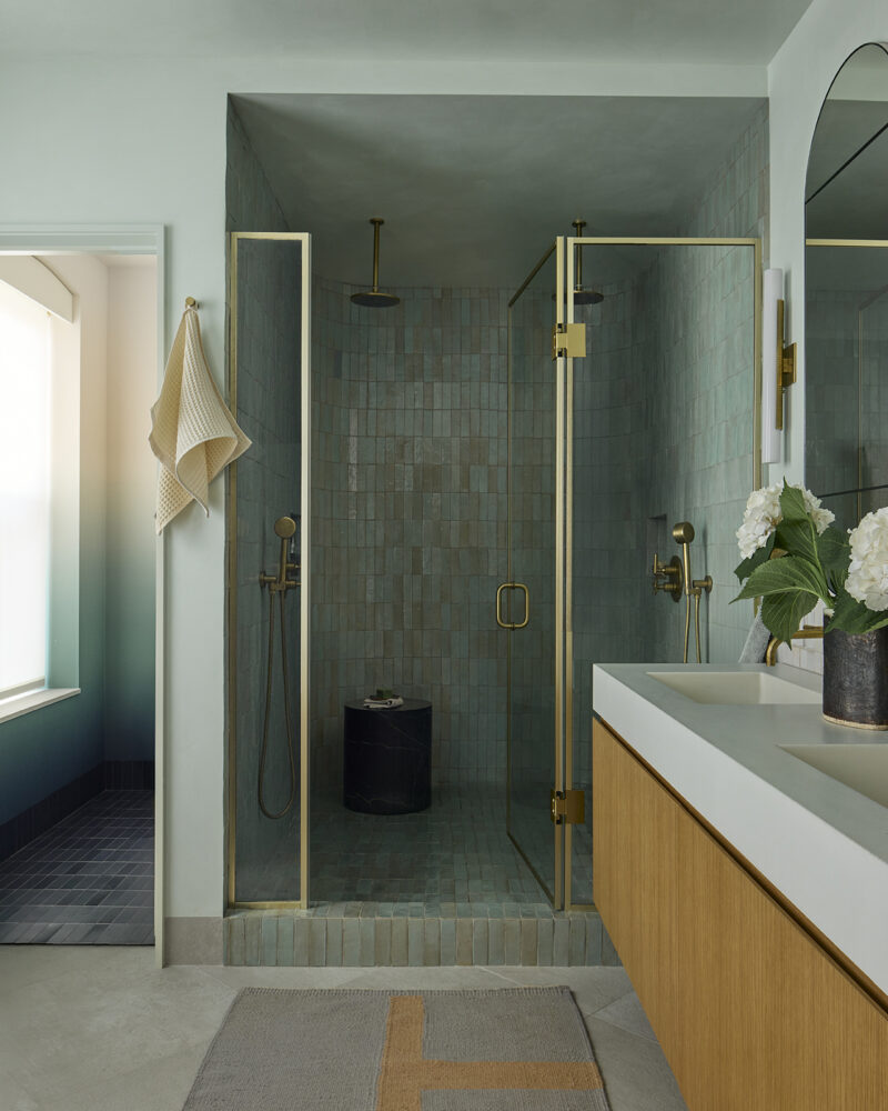 A walk-in shower with green tiled walls, a glass door, and gold fixtures, next to a double vanity with a wooden cabinet.