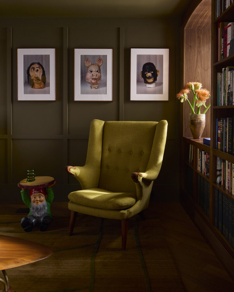A yellow-green wingback chair sits in a room with three framed masks on the wall, a gnome side table, and a built-in bookshelf.