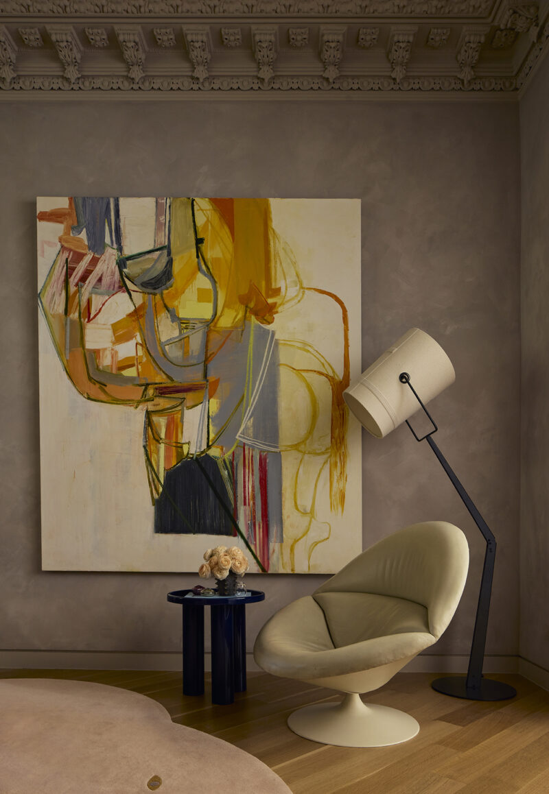 A large abstract painting with orange and yellow brushstrokes hangs behind a cream-colored swivel chair, a blue side table with a flower bouquet, and a floor lamp.