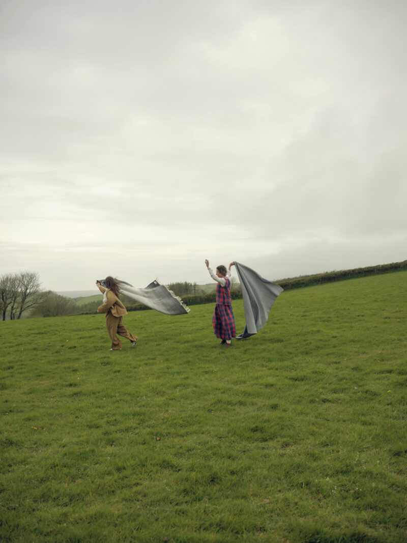 Two people walk on a grassy field holding blankets or fabric that flow behind them in the wind under a cloudy sky.