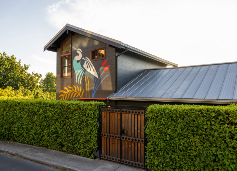 A dark-sided house at the Alki Lookout features a large wall mural of a blue heron surrounded by tall hedges and a wooden gate.