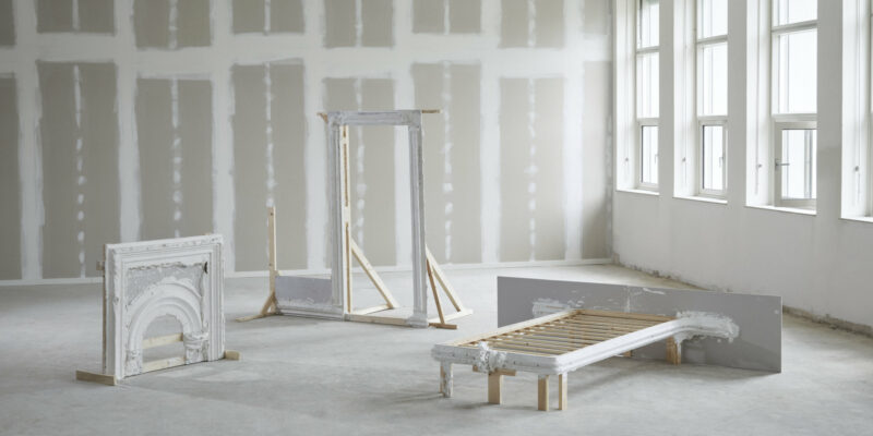 Three architectural fragments—a fireplace mantel, a door frame, and a bed frame—covered in white plaster, standing in an unfinished room with drywall and large windows.