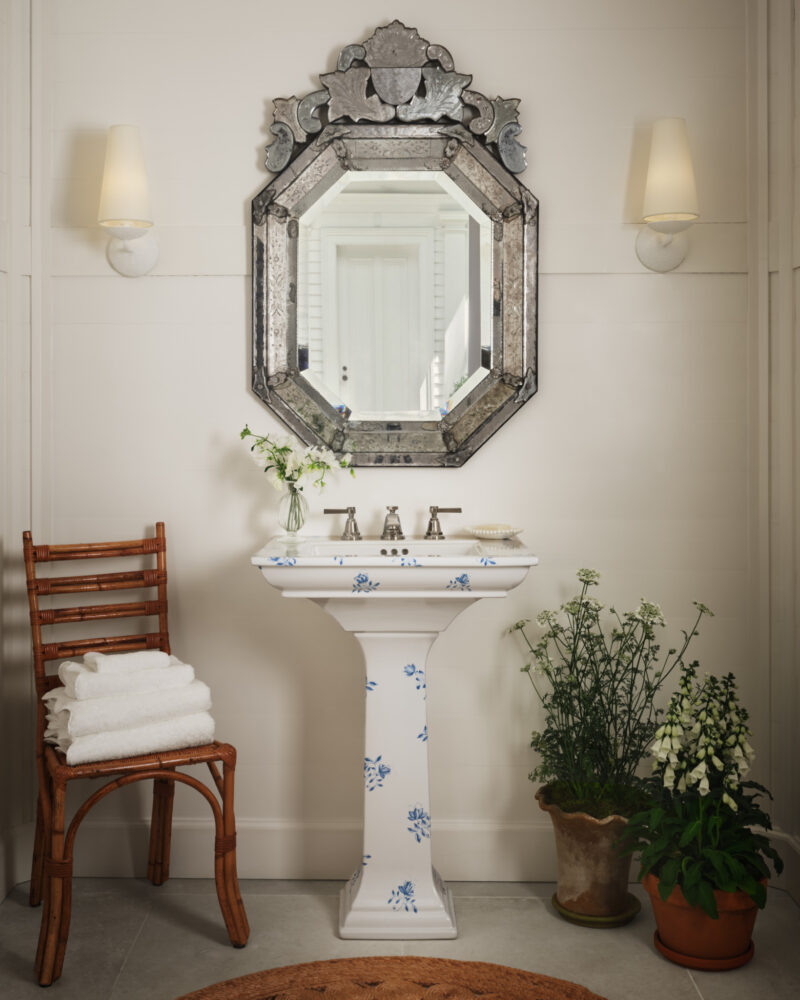 Pedestal sink with blue floral details, topped by an ornate octagonal mirror and flanked by a bamboo chair with stacked towels and potted plants.