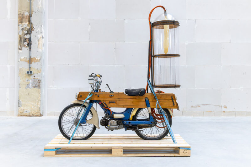 A vintage moped with wood-paneled sides and a tall, cylindrical glass container mounted on the back, standing on a wooden shipping pallet.