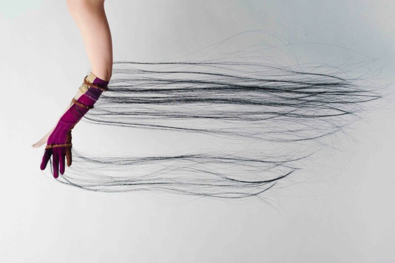 A gloved hand extending horizontally, with long, thin black strands trailing from the fingertips across a white background.