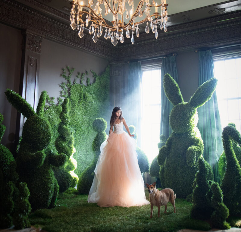 A girl in a long gown stands in an Adrien Broom-inspired room filled with large bunny-shaped topiaries and a small dog on a grass floor.
