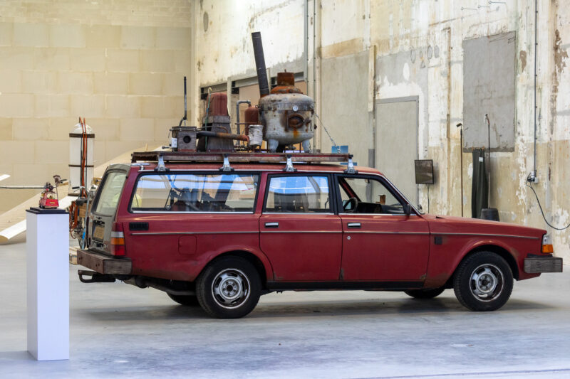 A weathered red station wagon with a custom metal furnace and piping mounted on its roof rack, displayed in an industrial gallery space.