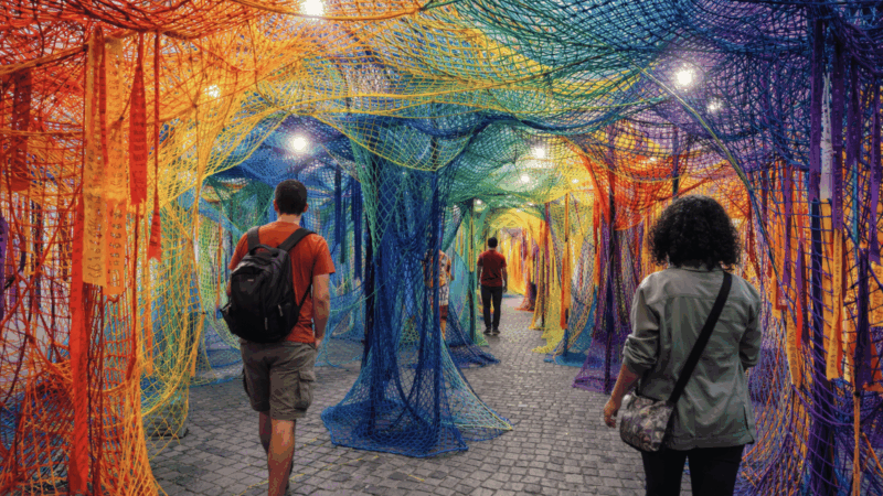 People walk through a path surrounded by a tunnel of multicolored, overlapping mesh nets with dangling ribbons.