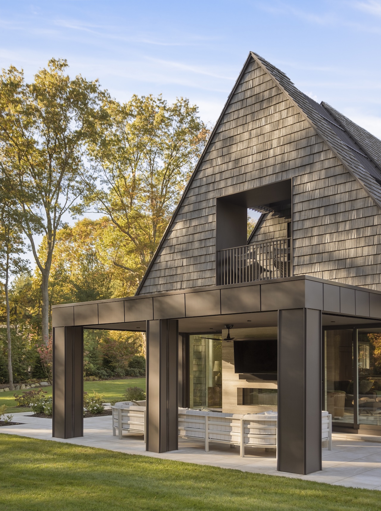 A modern house with a steep shingled roof, a recessed balcony, and a covered patio furnished with outdoor seating.