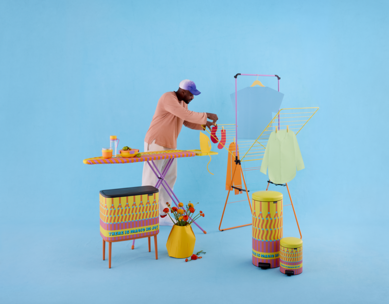 A person ironing a yellow sock on an ironing board next to a drying rack, decorative containers, and a vase of flowers.